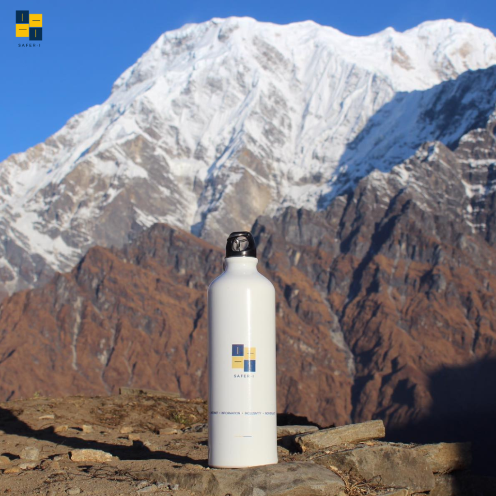 A bottle with the logo and mission of Safer-I printed on it before a mountain range showing the long way that Safer-I has to go