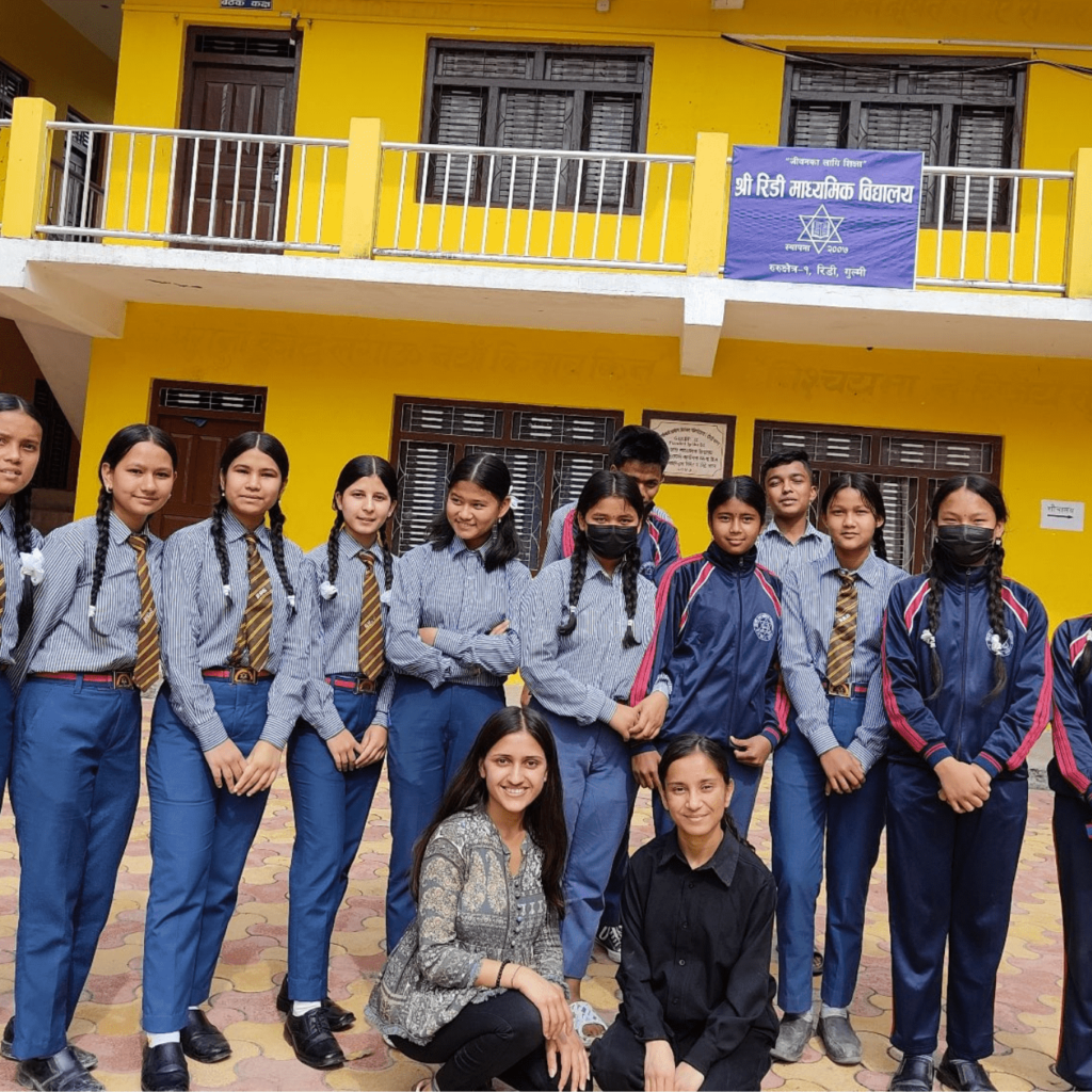 A group of people poses before a school with a board that reads ‘Ridi Higher Secondary School, Ridi, Gulmi’. Two boys in blue school uniforms are in the back row. In the middle row are 10 girls in the same blue school uniforms. Two women, one in a grey kurta and the other in a black shirt sit on their knees in the first row.