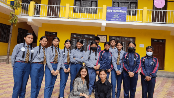 A group of people poses before a school with a board that reads ‘Ridi Higher Secondary School, Ridi, Gulmi’. Two boys in blue school uniforms are in the back row. In the middle row are 10 girls in the same blue school uniforms. Two women, one in a grey kurta and the other in a black shirt sit on their knees in the first row.