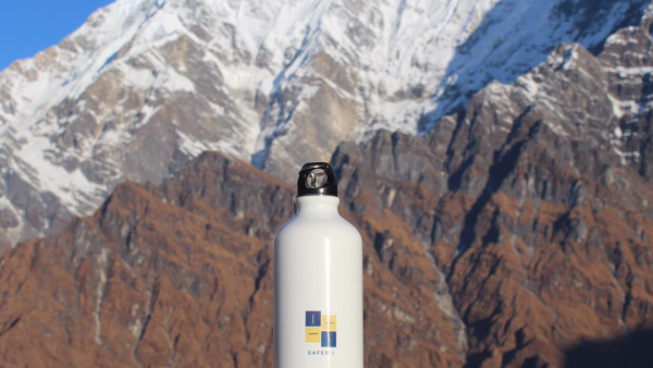 A bottle with the logo and mission of Safer-I printed on it before a mountain range showing the long way that Safer-I has to go